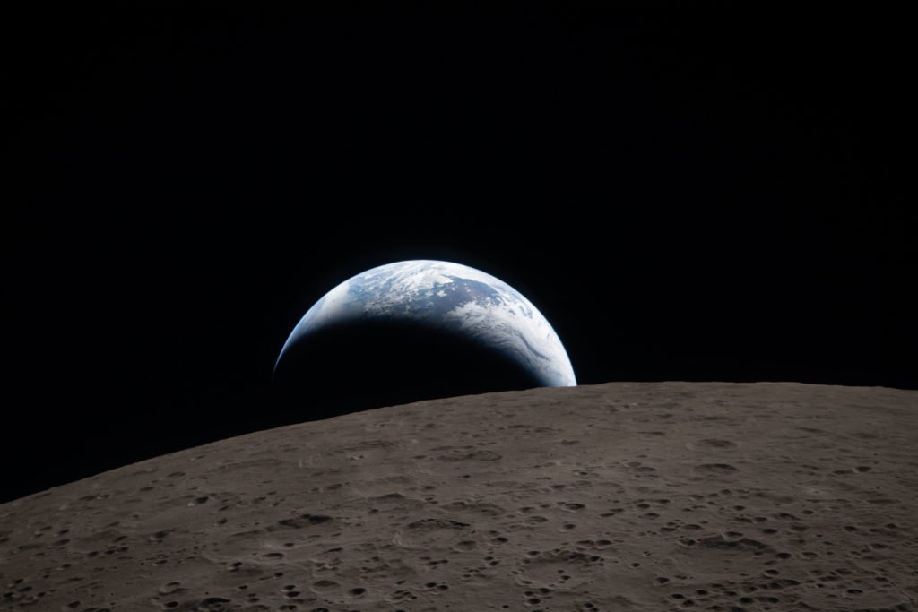 Earthset captured through the Orion spacecraft window at 6:41 p.m. EDT, April 6, 2026, during the Artemis II crew’s flyby of the Moon. A muted blue Earth with bright white clouds sets behind the cratered lunar surface. The dark portion of Earth is experiencing nighttime. On Earth’s day side, swirling clouds are visible over the Australia and Oceania region. In the foreground, Ohm crater has terraced edges and a flat floor interrupted by central peaks. Central peaks form in complex craters when the lunar surface, liquefied on impact, splashes upwards during the crater’s formation.   